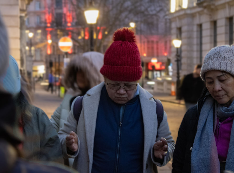 A lady wearing a red hat holds her hands out in prayer. She is surrounded by people in coats in soft streetlights.