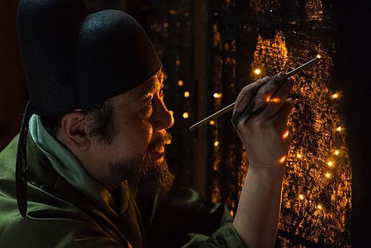 Yeong-Sil cubre con pintura negra los paneles de papel de las paredes del cuarto real y va creando las constelaciones, haciendo pequeñas perforaciones que se iluminan con la luz de la vela del chambelán que se encuentra al otro lado de la pared. | Lotte Entertainment