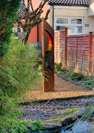 Stained glass outdoor sculpture with rainbow coloured wave in a pea gravel area with light shining through