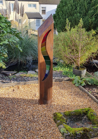 Stained glass outdoor sculpture with rainbow coloured wave in a pea gravel area