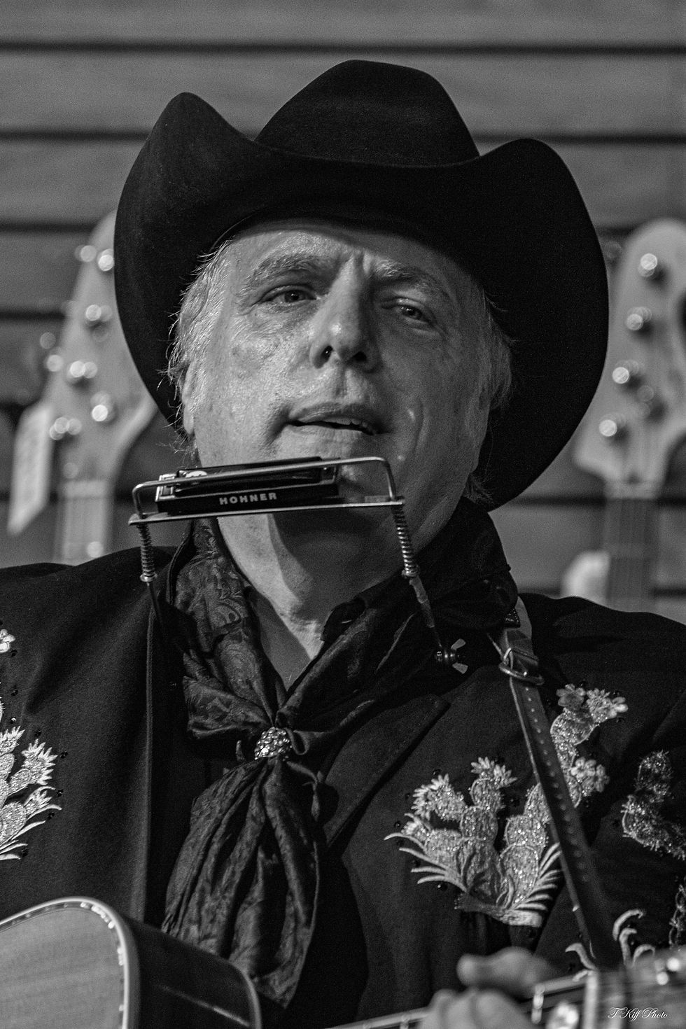 Man in cowboy hat plays guitar with harmonica. Wears patterned jacket, black and white setting. Background has blurred guitars.