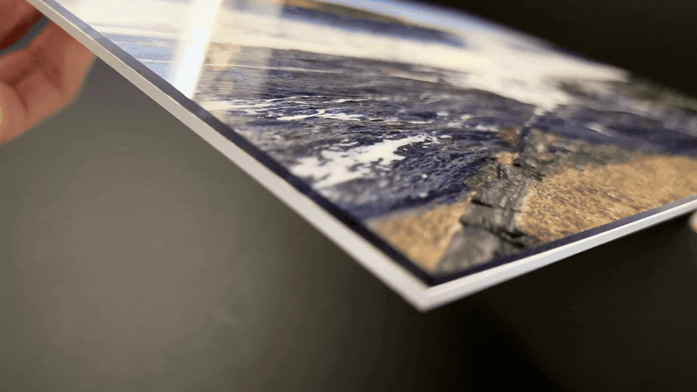 Hand holds a vibrant photo of a coastline with waves breaking on rocks. The glossy finish reflects light. Background is blurred.