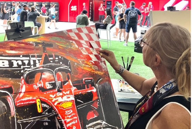 Anita Lewis painting a red Formula 1 car on canvas in a vibrant, public setting. People in the background, with red and black decor.