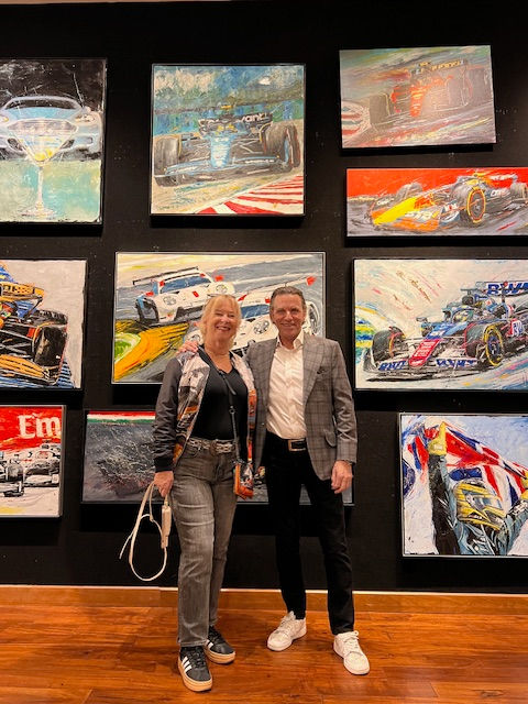 Two people standing in front of a wall displaying colorful racing car paintings. Wooden floor, both smiling, casual attire.