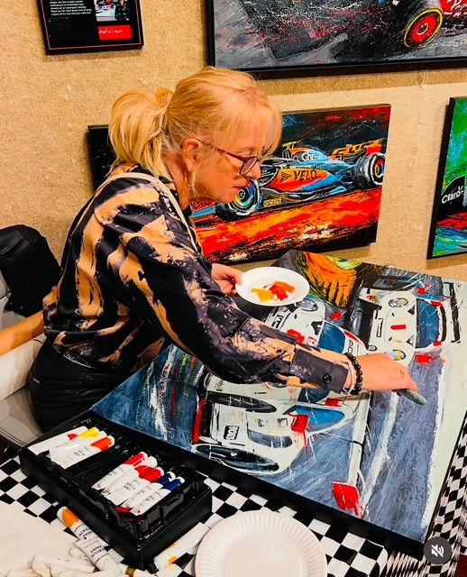 Anita Lewis painting race cars on canvas, surrounded by vibrant car artwork. Wearing a patterned shirt, using a palette with red and yellow paint.