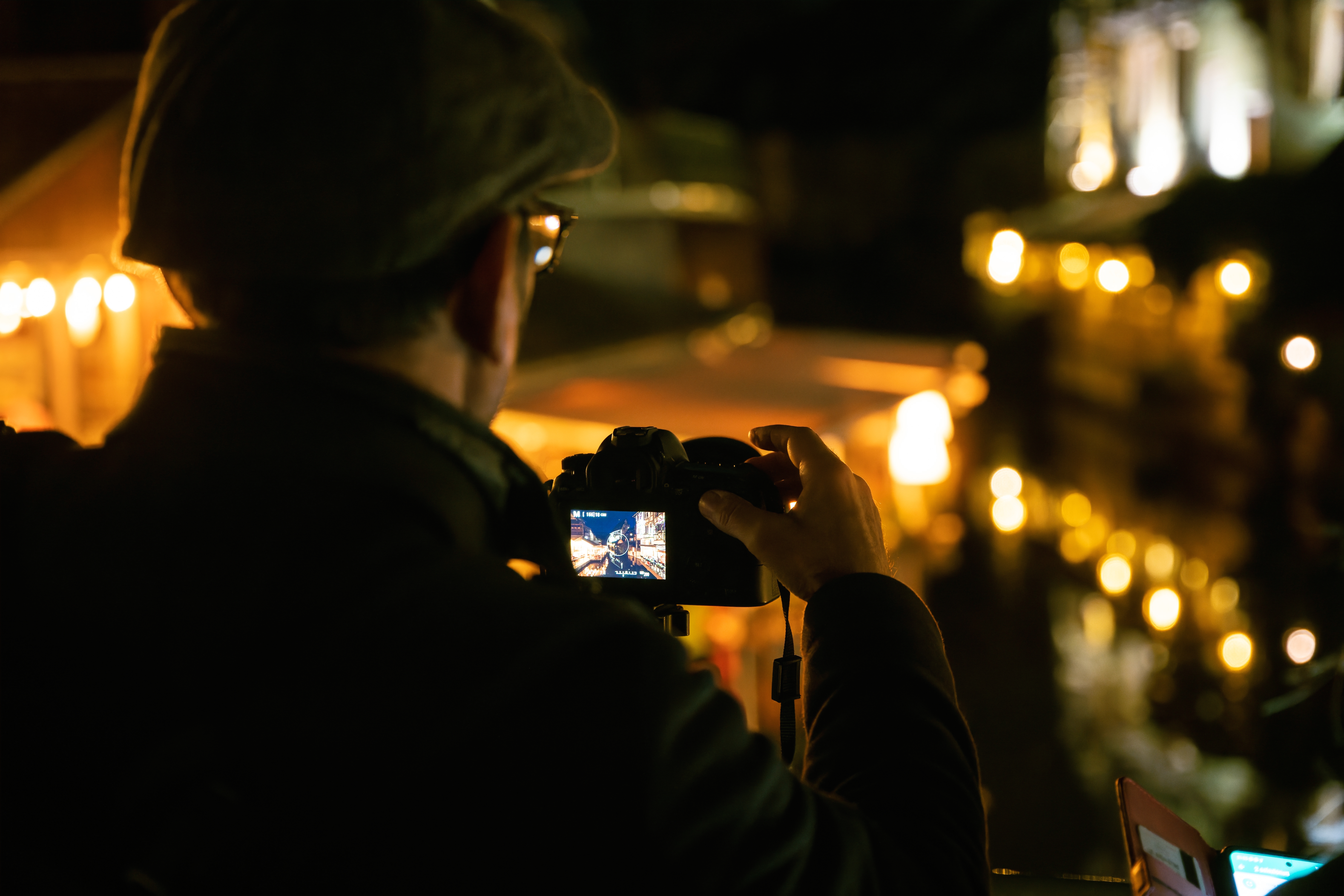 A person in a hat captures a night scene with a camera. Warm yellow lights and blurred background create a cozy ambiance.