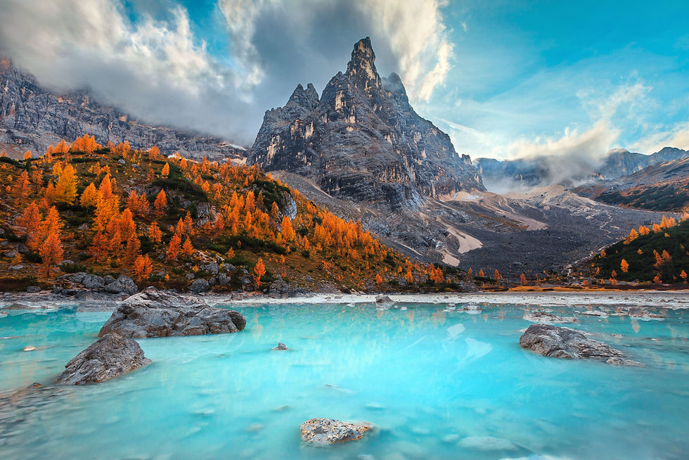 Turquoise lake with rocks, surrounded by orange autumn trees, below a towering mountain under a blue sky with clouds. Serene mood.