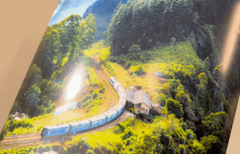 On glossy paper, a blue train curves through lush green hills with tall trees. A small building is nearby. Sunlight glistens, creating a serene, vibrant scene.