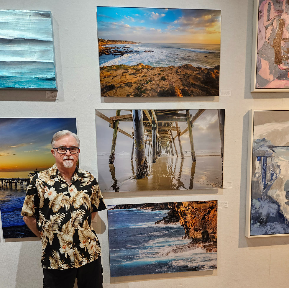 Richard Daugherty stands in art gallery, smiling. Coastal and pier photographs on white walls, wooden floor beneath. Relaxed mood.