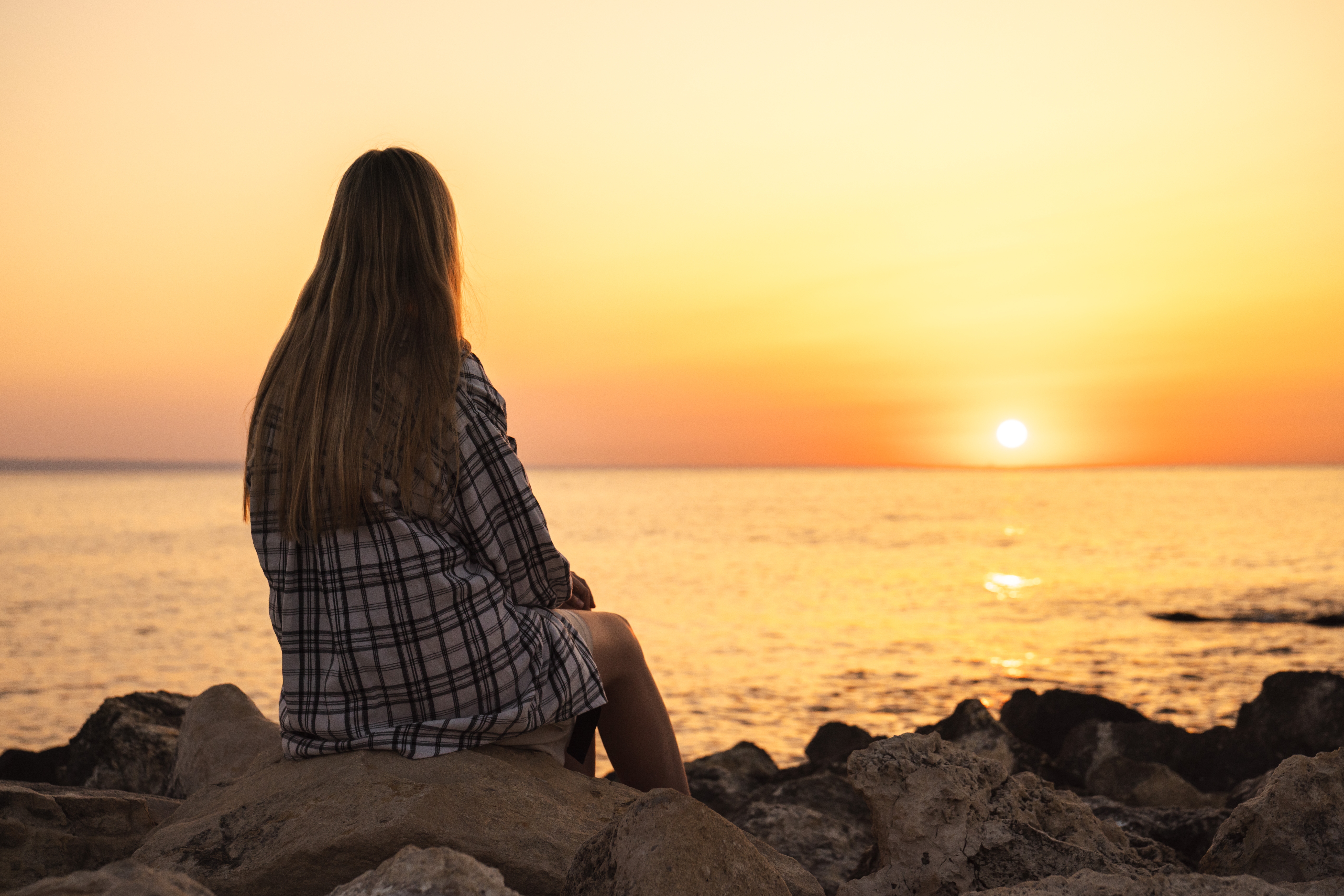 Woman in plaid shirt sits on rocky shore, watching a serene sunset over the ocean. Warm hues of orange and yellow fill the sky.