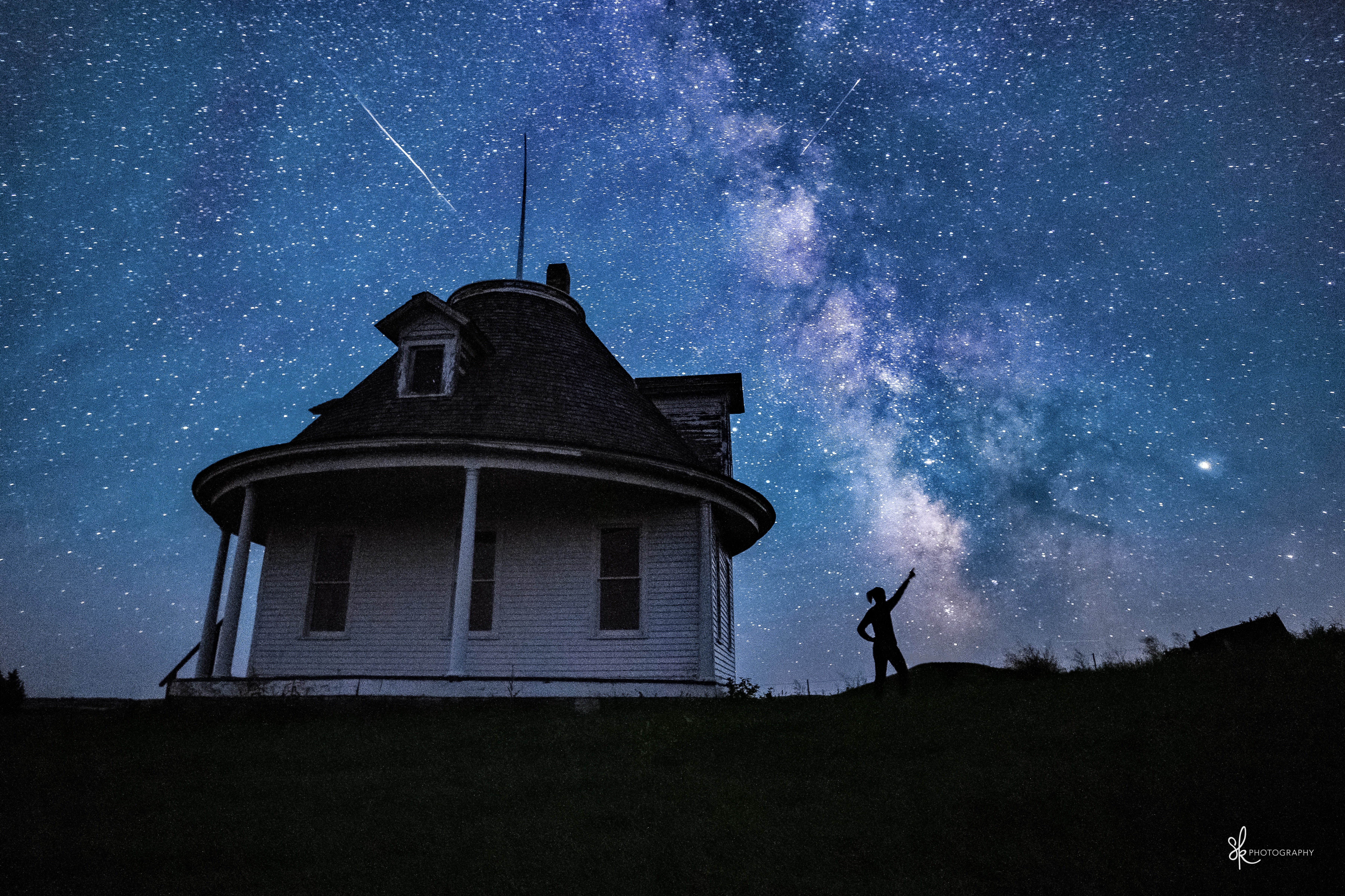 Silhouette of a person pointing at the starry night sky near a round house. Milky Way and shooting stars visible, creating a serene scene.