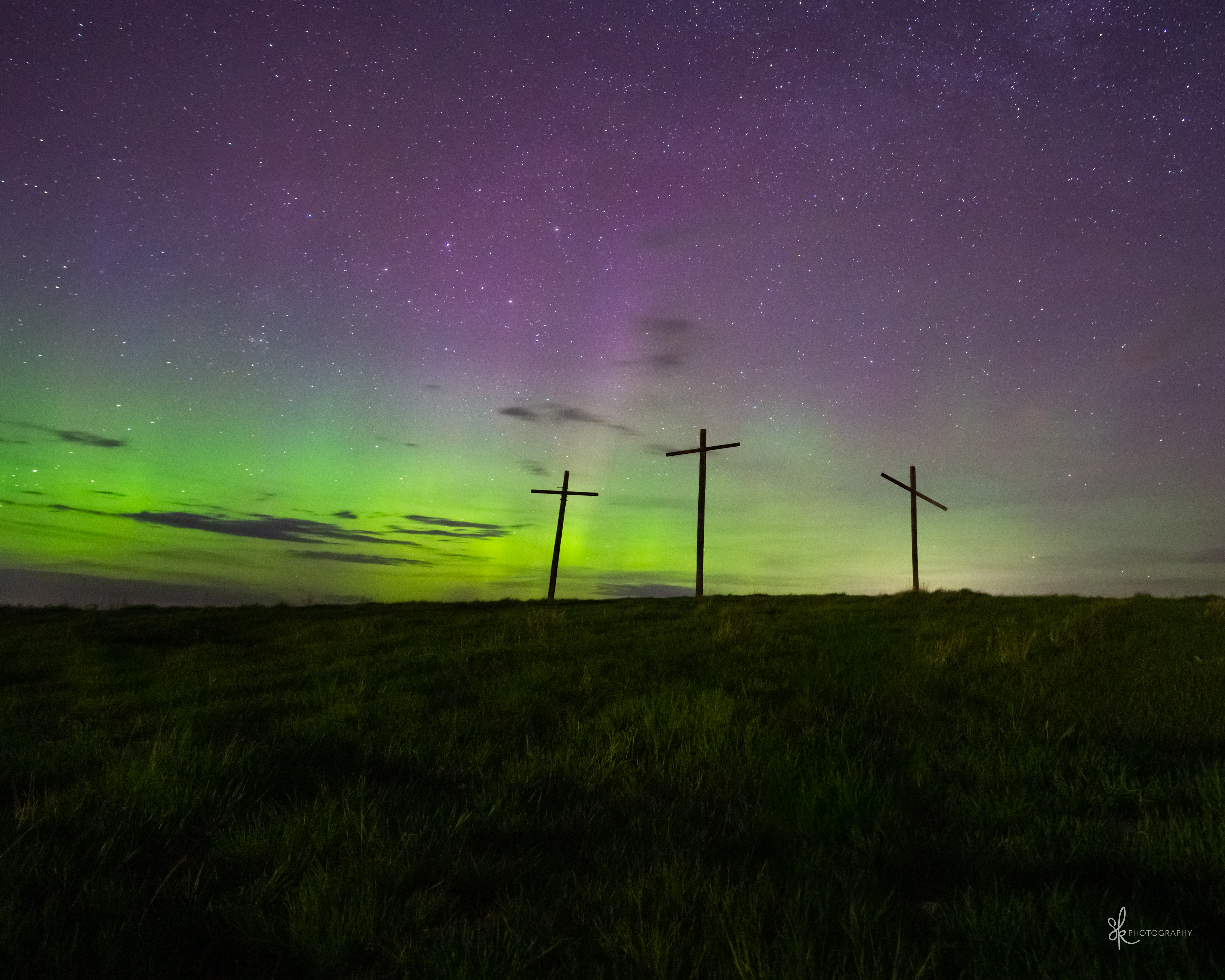 A dark field with aurora and stars in the night sky.  Three crosses stand tall in the middle-ground. 