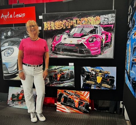 Anita Lewis in pink shirt smiling, standing beside vibrant car art display. Paintings show colorful race cars. Bold "Anita Lewis" sign above.