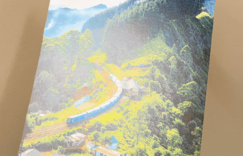 On fine art paper, a winding train moves through lush green hills under a bright sky, with vibrant colors evoking a serene, scenic atmosphere.