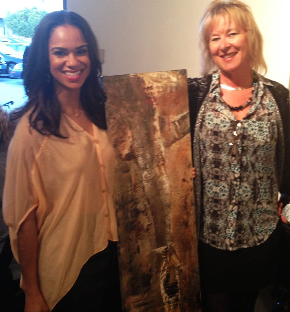 Two women smiling, holding an abstract painting in a gallery setting. One wears a beige blouse; the other a patterned shirt. Warm, casual mood.