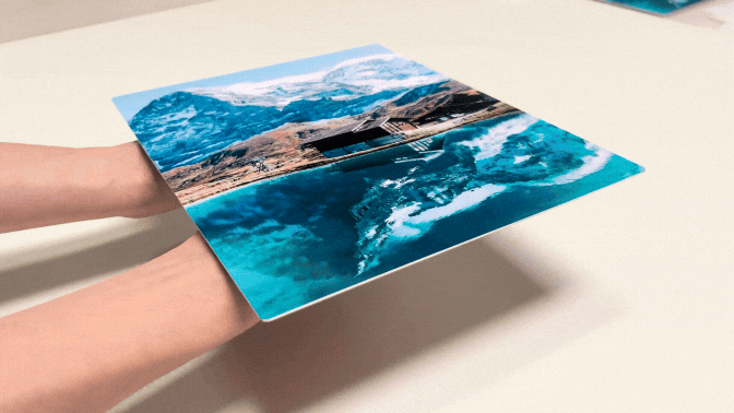 Hands hold a vivid HD Metal print on White Gloss of a mountain landscape and lake. The background is plain. The mood is serene and reflective.