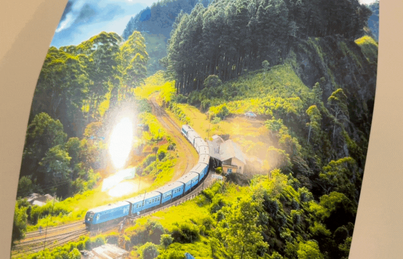 On metallic paper, a blue train winding through lush green hills under a bright sky, surrounded by dense tall trees; sunlight creates a serene atmosphere.