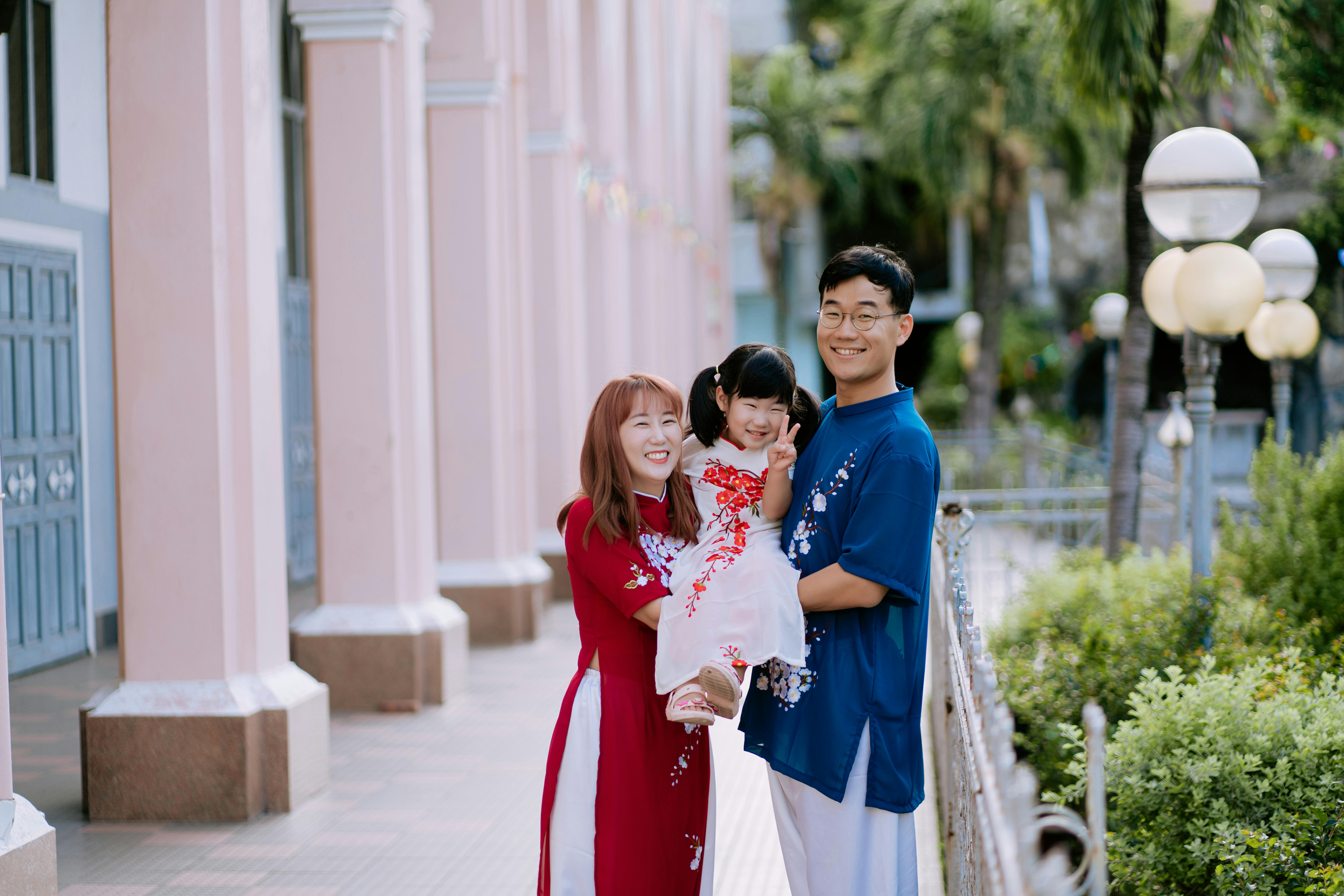 A family of three, parents in red and blue attire, joyfully hold their child in a white dress. Pink building and greenery in the background.