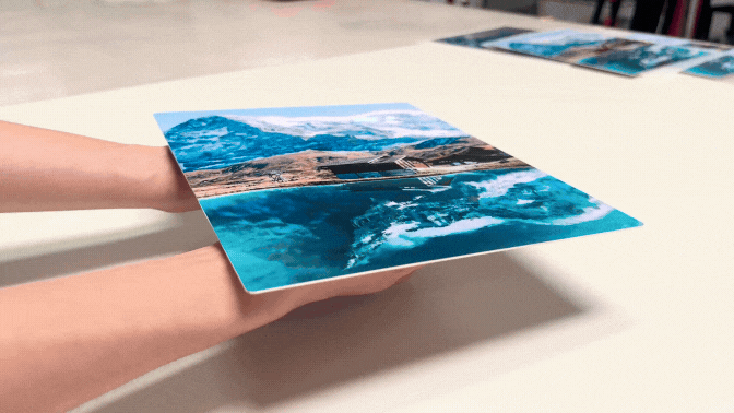 Hands hold a vivid HD Metal print on White Satin of a mountain landscape and lake. The background is plain. The mood is serene and reflective.