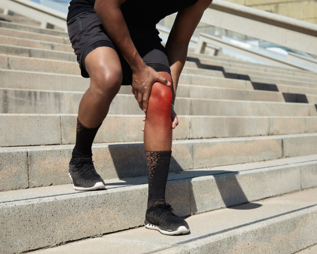 Person in athletic wear clutching a painful, red-inflamed knee on outdoor stairs, conveying discomfort and injury.