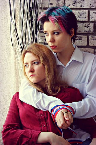 A couple of two women, with one with blue, pink and dark cropped hair and white shirt with her arms draped around another woman with long blonde hair wearing a red outfit. 