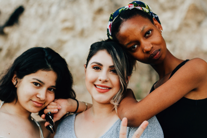 Two Latinx and one Black young woman standing together leaning on each other looking at the camera. 