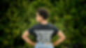 A Black woman with short dark hair and a large hoop earring looking over her left shoulder and her hands on her hips. We can read the back of her t-shirt "No homophobia. No violence. No racism. No sexism. Yes kindness. Yes peace