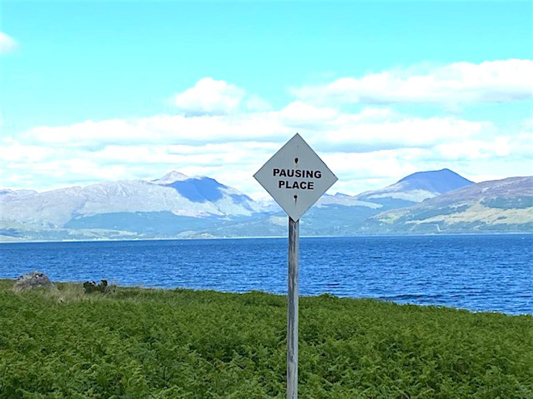 Thomas A Clark, Pausing Place. Photograph of a mountainous landscape in the distance, a body of water, grass in the foreground, and a roadsign (which is the artwork) that reads: PAUSING PLACE.