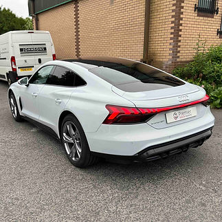 Rear three-quarter view of a white Audi e-tron GT coupé parked outside a brick building and a van