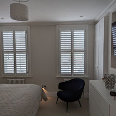 Cosy neutral bedroom with two tall windows featuring white plantation shutters and a dark blue chair