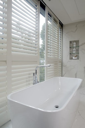 Freestanding bathtub and tap next to a white marbled wall and windows fitted with plantation shutters