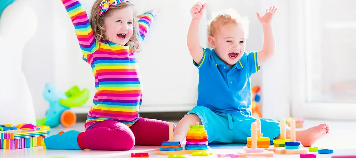 Two happy children in bright clothing playing with building blocks and raising their arms in a sunny playroom