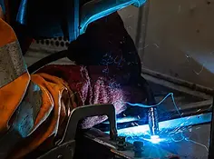 A person welds metal, surrounded by bright blue sparks and intense light