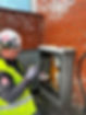 Worker in yellow high-visibility vest and hard hat inspecting outdoor electrical cabinet on a red brick wall