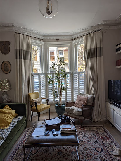 Bay window with white cafe style shutters, full-length curtains, and a tall potted plant in a living room