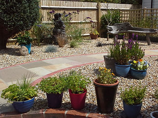 A peaceful garden with a gravel path, various potted plants, a wooden bench, and a small fountain