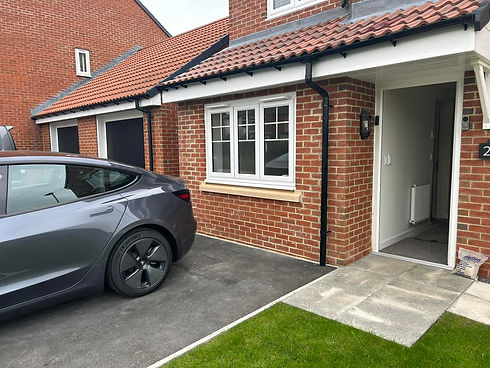 A modern gray car is parked in the driveway of a brick house with an open front door