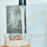 A hand holds a clear glass cup labeled "Quooker" under a running faucet, filling it with water