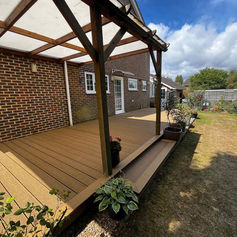 Large rear garden composite decking with a timber pergola structure and clear roofing panel on a sunny day