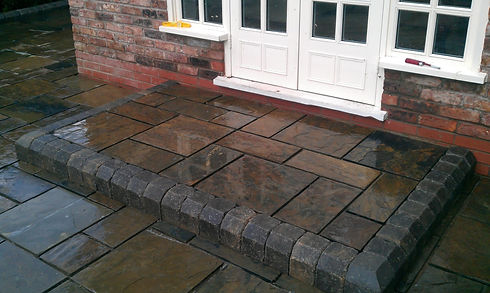 Wet stone patio with dark grey tiles and a brick border, leading to a white wooden door on a brick house