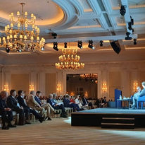 An indoor conference hall with a chandelier and people seated watching a stage.