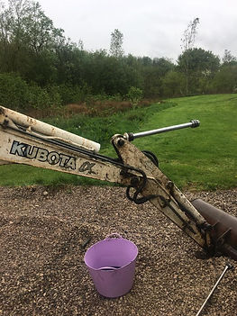 A backhoe arm labeled "KUBOTA" extends over gravel, with a grassy field and trees in the background