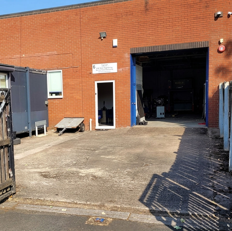 Red brick industrial unit exterior with open roller shutter door and paved driveway yard area
