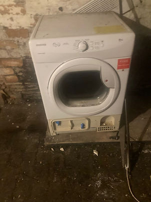 An old, white tumble dryer or washing machine with the front panel missing against a dark brick wall