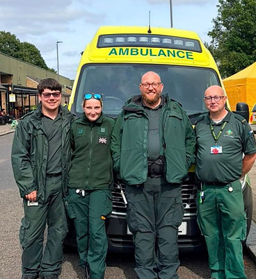 Four paramedic crew members in green uniforms posing smiling in front of an ambulance.