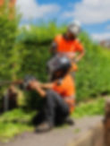 Two workers in orange safety gear and helmets trim tall hedges with power tools