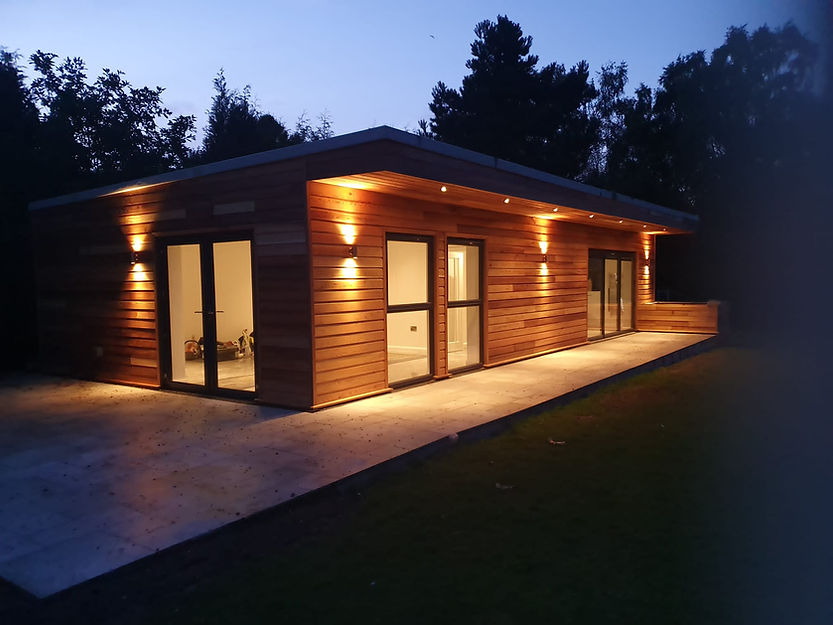 Modern timber-clad garden room or office at dusk with exterior down lighting illuminating the patio