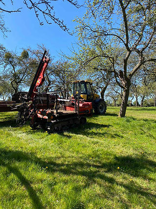 A red and black drilling rig and a yellow tractor are parked in a grassy area