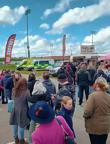 A large crowd of people standing outdoors near food trucks and ambulances under a blue sky