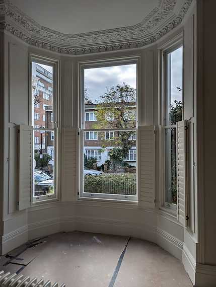 Close-up of a bay window with white plantation shutters partially folded back to show the street outside